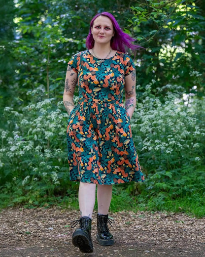 A model with bright purple hair stands outdoors in a wooded area wearing a cap-sleeve knee-length dress with a colourful print on a black base featuring squirrels among autumn foliage and pine cones.