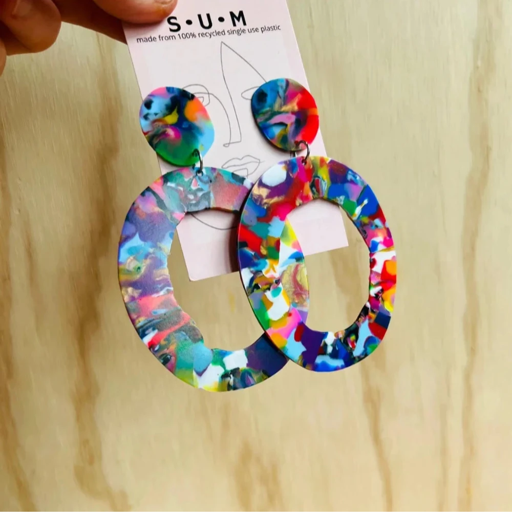 Colorful earrings held by a hand with a visible brand label on a beige background