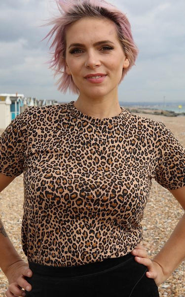 Image is of a person standing on a beach, with their hands on their hips, wearing a leopard print t shirt.