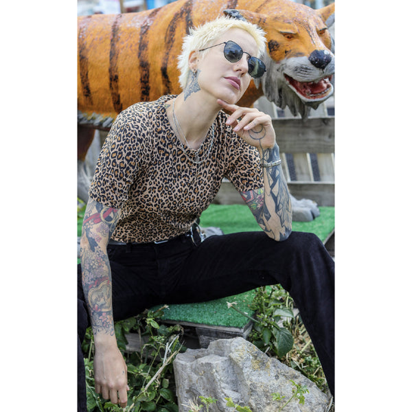 Image is of a person sitting with their hand under their chin, wearing a leopard print tee and black pants.