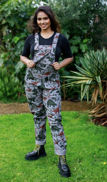 Person standing in a garden, wearing grey dungarees with a dinosaur print.