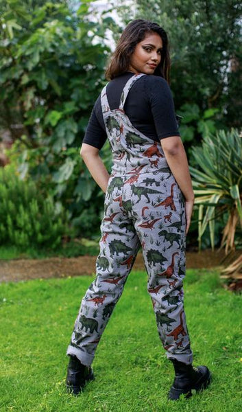 Person standing in a garden, looking back over their shoulder, wearing grey dungarees with a dinosaur print.