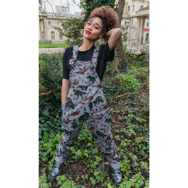 Person standing in a garden, wearing grey dungarees with a dinosaur print.